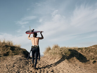 Mann, der auf einem Pfad in den Sanddünen in Richtung Meer geht und ein Surfbrett auf dem Kopf balanciert - MINF09402