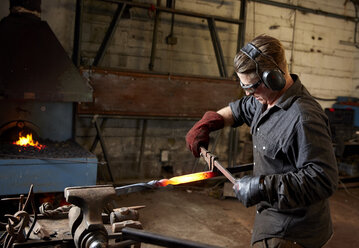 Handwerklicher Metallarbeiter mit Gehörschutz und Schutzbrille bei der Arbeit mit Hammer und Amboss, um ein glühendes Metallstück mit einer Drehung zu formen. - MINF09405