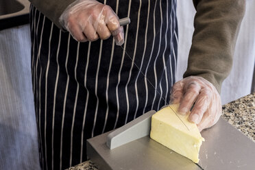 High angle close up of man wearing apron standing at counter in a delicatessen, slicing cheese with wire cutter. - MINF09491