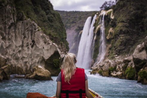 Rückansicht einer Frau, die in einem Boot auf einem Fluss vor einem Wasserfall sitzt - CAVF53192