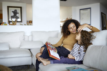 Mutter liest Bilderbuch für süße Tochter, während sie zu Hause auf dem Sofa sitzt - CAVF53214