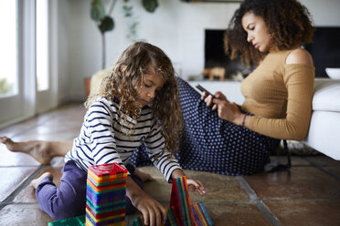 Mutter, die ein Mobiltelefon benutzt, während ihre Tochter im Wohnzimmer mit Spielzeug spielt - CAVF53218