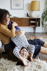 Happy mother holding daughter upside down while sitting on floor at home - CAVF53219