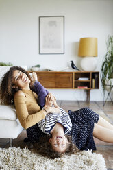 Happy mother holding daughter upside down while sitting on floor by sofa at home - CAVF53220