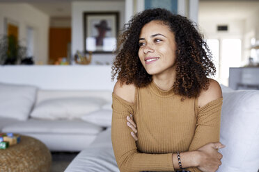 Happy woman with arms crossed looking away while sitting on sofa at home - CAVF53227