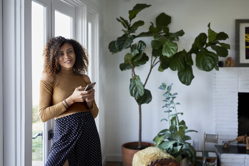 Portrait of smiling woman using mobile phone while standing by window at home - CAVF53228
