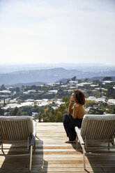 Side view of woman talking on mobile phone while sitting on deckchair against sky - CAVF53230