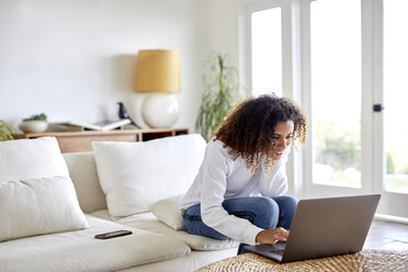 Lächelnde Frau, die einen Laptop benutzt, während sie zu Hause auf dem Sofa sitzt - CAVF53252