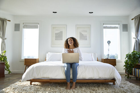 Glückliche Frau, die einen Laptop benutzt, während sie zu Hause auf dem Bett sitzt - CAVF53254