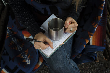 Mittelteil einer Frau, die einen leeren Becher hält und mit einem Stift in ihr Tagebuch schreibt - CAVF53279