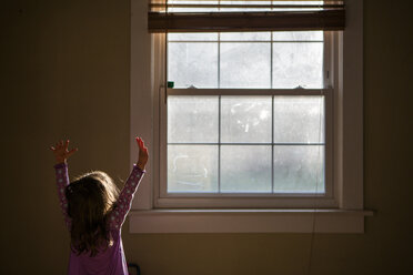 Girl with arms raised standing by window at home - CAVF53289