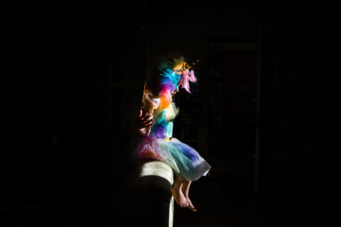 Side view of girl wearing colorful unicorn costume sitting on sofa in darkroom at home - CAVF53296