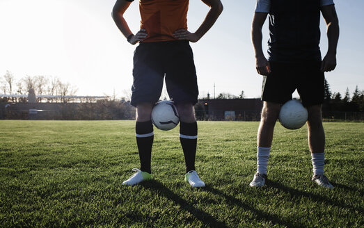 Low section of male friends with soccer balls standing on grassy field against clear sky during sunset - CAVF53356