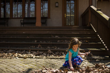 Mädchen sammelt trockene Blätter, die im Herbst auf dem Fußweg vor dem Haus liegen - CAVF53364