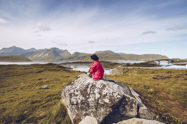 Norway, Lofoten, man wearing red rainjacket sitting on a rock looking at view - RSGF00054