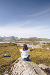 Norway, Lofoten, back view of young woman sitting on a rock doing yoga - RSGF00056