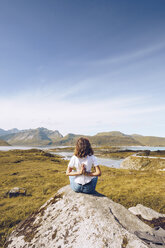 Norway, Lofoten, back view of young woman sitting on a rock doing yoga exercise - RSGF00057