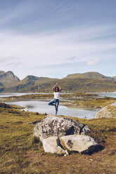 Norwegen, Lofoten, junge Frau steht mit einem Bein auf einem Felsen und macht Yoga - RSGF00058