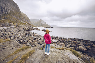 Norwegen, Lofoten, Rückenansicht einer jungen Frau in Regenjacke, die auf einem Felsen steht und in die Ferne schaut - RSGF00064