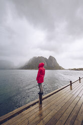 Norwegen, Lofoten, Hamnoy, Mann mit roter Regenjacke steht auf Holzpfahl und schaut in die Ferne - RSGF00072