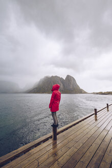 Norwegen, Lofoten, Hamnoy, Mann mit roter Regenjacke steht auf Holzpfahl und schaut in die Ferne - RSGF00072