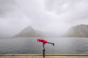 Norway, Lofoten, Hamnoy, man wearing red rain jacket balancing on wooden stake - RSGF00073
