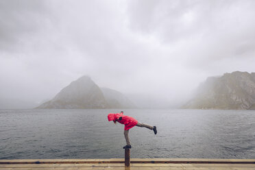 Norwegen, Lofoten, Hamnoy, Mann mit roter Regenjacke balanciert auf Holzpfahl - RSGF00073