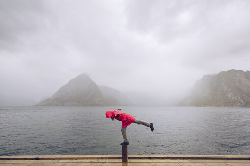 Norwegen, Lofoten, Hamnoy, Mann mit roter Regenjacke balanciert auf Holzpfahl - RSGF00073