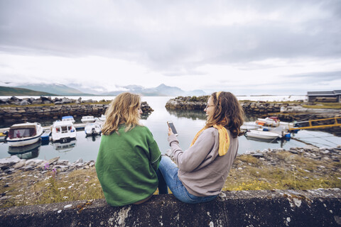 Norwegen, Senja, zwei junge Frauen sitzen an einem kleinen Hafen mit Mobiltelefon, lizenzfreies Stockfoto