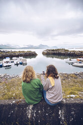 Norwegen, Senja, zwei junge Frauen sitzen an einem kleinen Hafen mit Mobiltelefon - RSGF00076