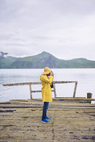 Norwegen, Senja, Mann steht auf einem baufälligen Steg an der Küste und macht ein Foto, lizenzfreies Stockfoto