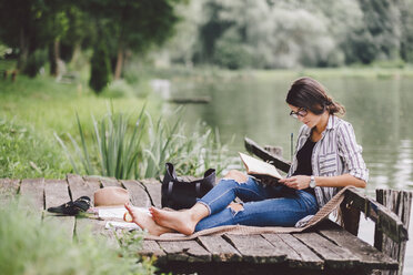 Frau liest ein Buch, während sie auf einem Steg am See im Wald sitzt - CAVF53393