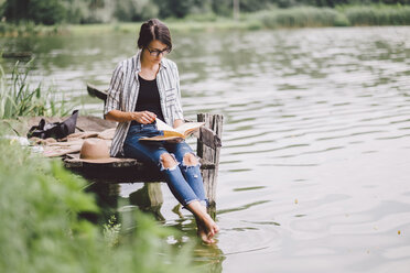 Frau liest ein Buch, während sie auf einem Steg über einem See im Wald sitzt - CAVF53396