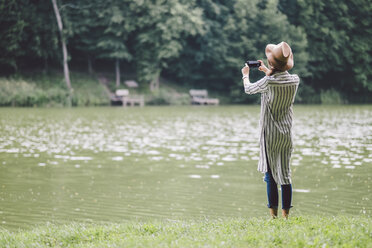 Rear view of woman photographing with mobile phone while standing at lakeshore in forest - CAVF53400