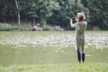 Rückansicht einer Frau, die mit ihrem Handy fotografiert, während sie am Seeufer im Wald steht - CAVF53400