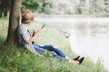 Frau benutzt Mobiltelefon, während sie auf einer Wiese am Seeufer im Wald sitzt - CAVF53401
