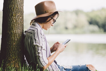 Woman using smart phone while sitting by tree against lake in forest - CAVF53402