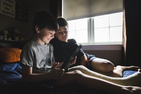 Smiling boy with broken leg using tablet computer while sitting by brother on bed at home - CAVF53411