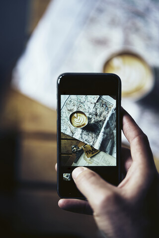 Ausgeschnittene Hand eines Mannes, der eine Kaffeetasse mit einer Karte auf einem Tisch mit einem Mobiltelefon zu Hause fotografiert, lizenzfreies Stockfoto