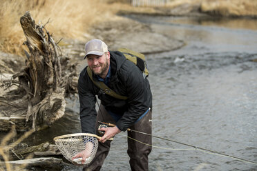 Lächelnder Mann, der im Fluss stehend einen Fisch aus einem Schmetterlingsfangnetz entfernt - CAVF53512