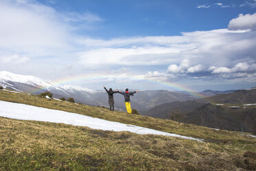 Rear view of friends with arms outstretched standing against mountains and rainbow during winter - CAVF53520