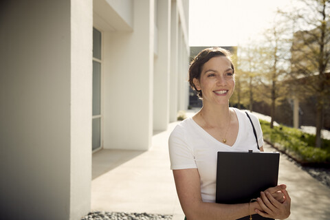 Smiling woman holding laptop computer while standing by building in city stock photo