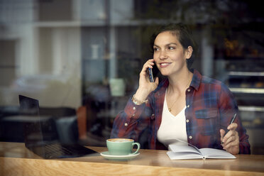 Frau, die in einem Café sitzt und mit ihrem Handy telefoniert, gesehen durch ein Fenster - CAVF53529