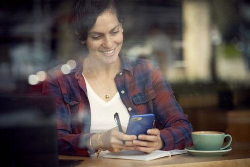 Lächelnde Frau mit Kaffee und Buch auf dem Tisch, die ein Mobiltelefon in einem Café benutzt, gesehen durch ein Fenster - CAVF53531