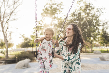 Happy mother pushing cute daughter swinging at playground - CAVF53545