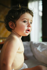 Side view of curious shirtless baby girl sitting on bed at home - CAVF53642