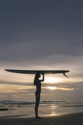 Seitenansicht einer jungen Frau, die ein Surfbrett auf dem Kopf trägt, während sie am Strand gegen den Himmel bei Sonnenuntergang steht - CAVF53671