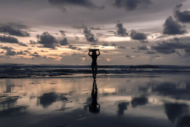 Silhouette young woman carrying surfboard on head at beach against cloudy sky during sunset - CAVF53673