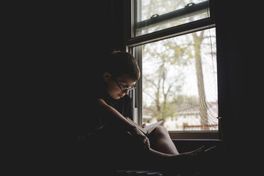 Boy reading book while sitting by window in darkroom at home - CAVF53679