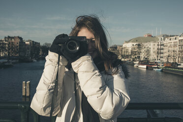 Lächelnde junge Frau beim Fotografieren mit Kamera am Kanal in der Stadt - CAVF53691